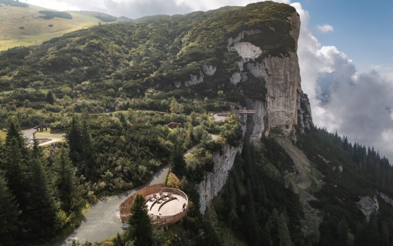 Eine runde hölzerne Aussichtsplattform überblickt einen steilen, bewaldeten Berghang mit einer felsigen Steilwand und einem wolkenverhangenen Himmel im Hintergrund.