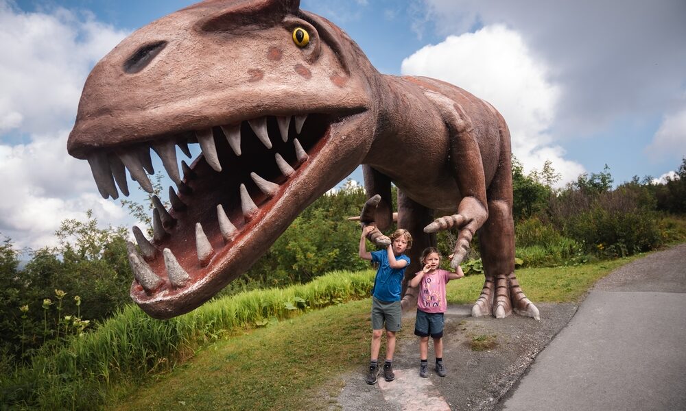 Zwei Kinder stehen vor einer großen, realistischen Dinosaurierstatue mit offenem Maul und scharfen Zähnen in einem Park im Freien.