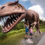 Zwei Kinder stehen vor einer großen, realistischen Dinosaurierstatue mit offenem Maul und scharfen Zähnen in einem Park im Freien.
