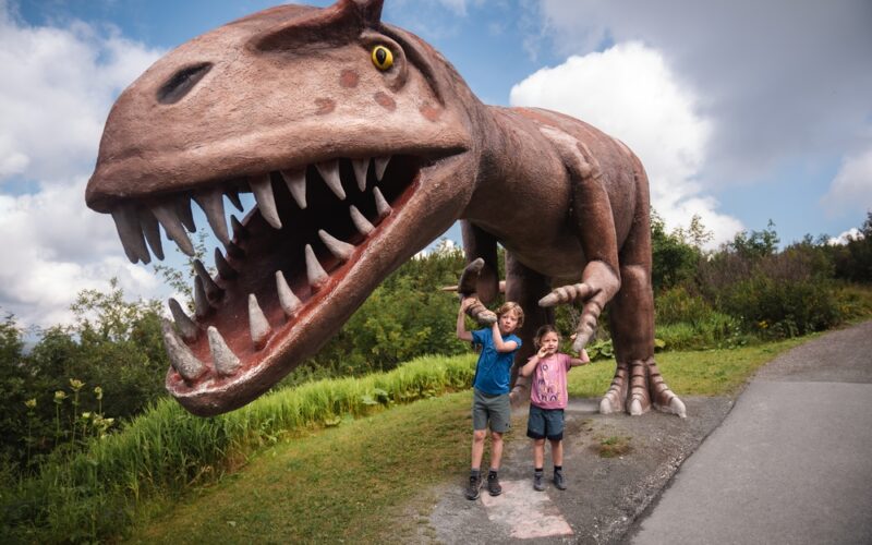 Zwei Kinder stehen vor einer großen, realistischen Dinosaurierstatue mit offenem Maul und scharfen Zähnen in einem Park im Freien.