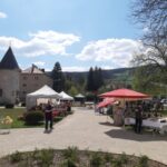 Markt im Freien mit Ständen und Menschen an einem sonnigen Tag, umgeben von Gebäuden, Bäumen und Hügeln im Hintergrund.
