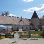 Open-Air-Markt mit mehreren Ständen und Zelten auf einem Stadtplatz, mit einem Kirchturm und historischen Gebäuden im Hintergrund unter einem teilweise bewölkten Himmel.