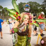 Frau in einem grünen Feenkostüm mit Flügeln und Blumen bläst Seifenblasen bei einer Veranstaltung im Freien, mit Menschen, die im Hintergrund spazieren gehen.