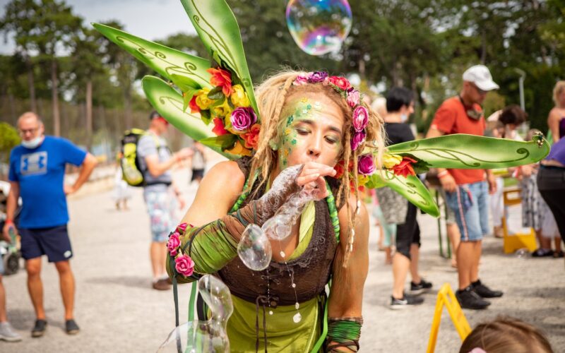Frau in einem grünen Feenkostüm mit Flügeln und Blumen bläst Seifenblasen bei einer Veranstaltung im Freien, mit Menschen, die im Hintergrund spazieren gehen.