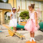 Zwei junge Mädchen, die als Feen verkleidet sind, spielen draußen; die eine balanciert auf einer hölzernen Wippe, während die andere daneben steht und zuschaut.