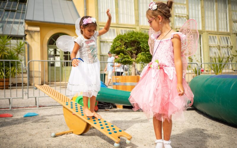 Zwei junge Mädchen, die als Feen verkleidet sind, spielen draußen; die eine balanciert auf einer hölzernen Wippe, während die andere daneben steht und zuschaut.