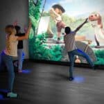 Drei Kinder interagieren mit einer auf eine Wand projizierten animierten Szene, die einen Cartoon-Forscher und einen Dinosaurier in einer Dschungelumgebung zeigt.