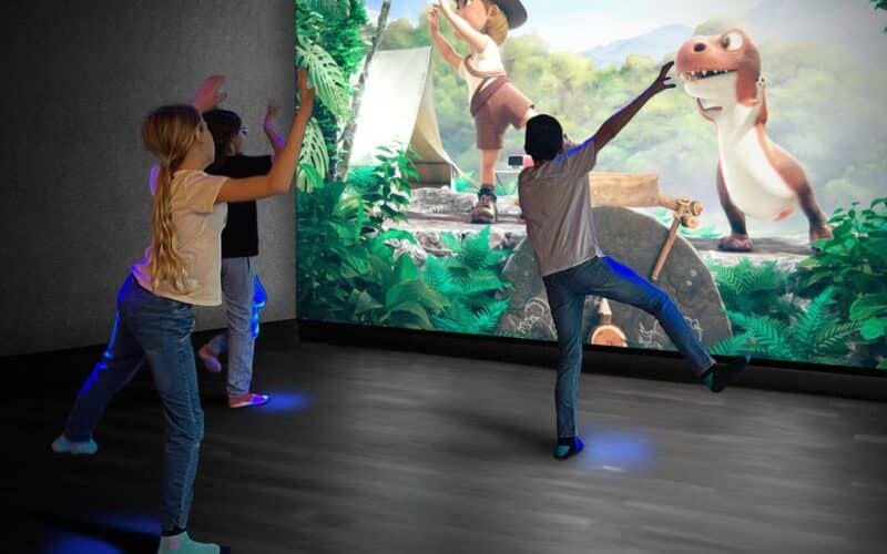 Drei Kinder interagieren mit einer auf eine Wand projizierten animierten Szene, die einen Cartoon-Forscher und einen Dinosaurier in einer Dschungelumgebung zeigt.