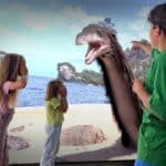 Drei Kinder stehen an einem Strand, schauen überrascht und zeigen auf einen großen, lebhaften prähistorischen Vogel, der in der Nähe des Ufers auftaucht.