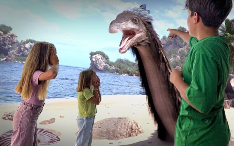 Drei Kinder stehen an einem Strand, schauen überrascht und zeigen auf einen großen, lebhaften prähistorischen Vogel, der in der Nähe des Ufers auftaucht.