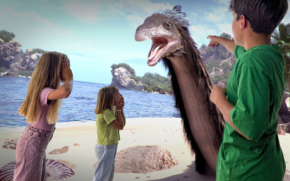Drei Kinder stehen an einem Strand, schauen überrascht und zeigen auf einen großen, lebhaften prähistorischen Vogel, der in der Nähe des Ufers auftaucht.