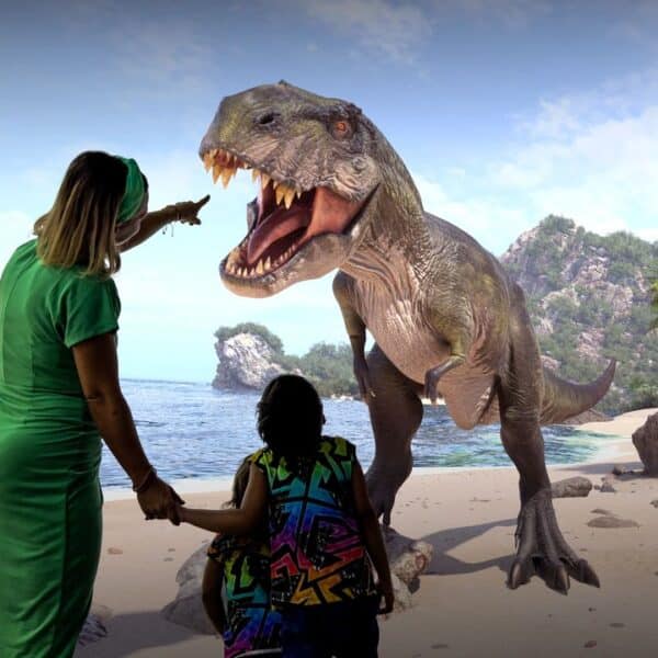 Zwei Menschen stehen an einem Strand, einer von ihnen zeigt auf ein großes, lebensechtes Dinosauriermodell vor ihnen, mit Felsen, Bäumen und Wasser im Hintergrund.