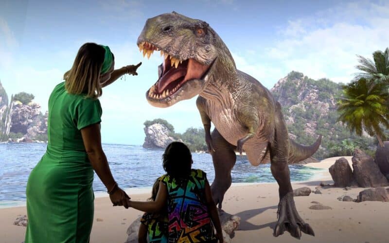 Zwei Menschen stehen an einem Strand, einer von ihnen zeigt auf ein großes, lebensechtes Dinosauriermodell vor ihnen, mit Felsen, Bäumen und Wasser im Hintergrund.