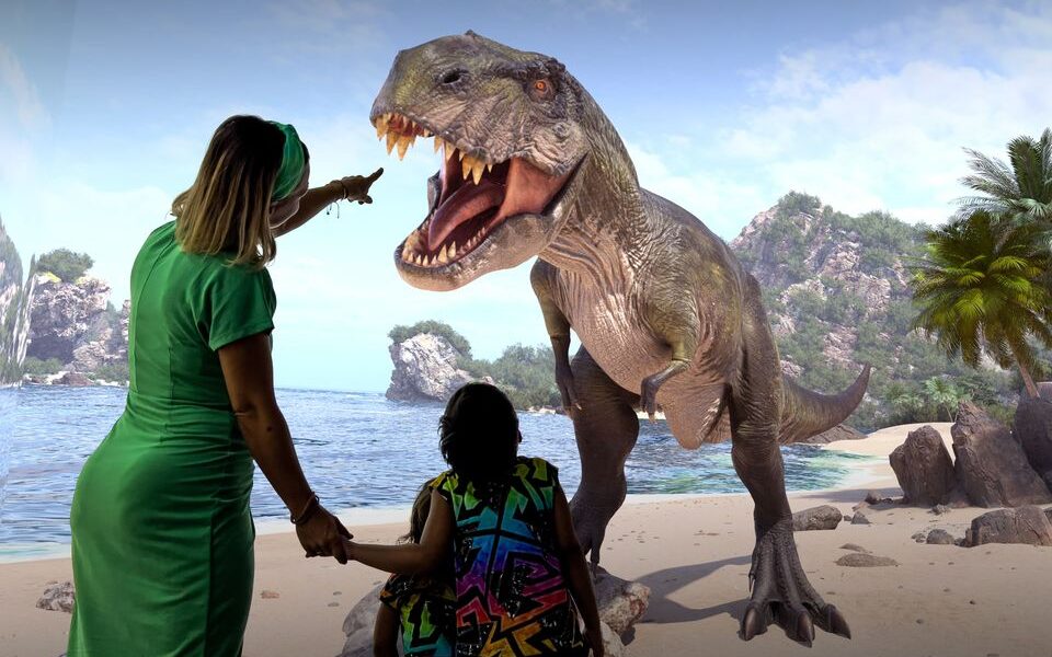Zwei Menschen stehen an einem Strand, einer von ihnen zeigt auf ein großes, lebensechtes Dinosauriermodell vor ihnen, mit Felsen, Bäumen und Wasser im Hintergrund.