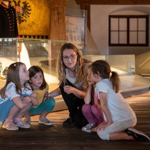 Eine Frau hockt mit drei jungen Mädchen in einem Museum, in dem im Hintergrund historische Ausstellungsstücke und Artefakte zu sehen sind.