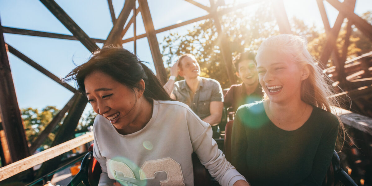 Vier Personen fahren tagsüber in einer Achterbahn und lächeln und lachen, während das Sonnenlicht durch die Holzkonstruktion im Hintergrund scheint.