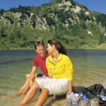 Ein Mann und eine Frau sitzen am Ufer eines Sees, die Füße im Wasser, Wanderschuhe und ein Rucksack in der Nähe, im Hintergrund ein grasbewachsener Berg.