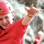 Ein Kind, das einen roten Helm und ein rotes Hemd trägt, lächelt und macht eine Geste mit einer Hand, während es im Freien klettert; eine andere Person ist unscharf im Hintergrund zu sehen.