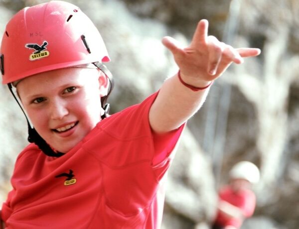 Ein Kind, das einen roten Helm und ein rotes Hemd trägt, lächelt und macht eine Geste mit einer Hand, während es im Freien klettert; eine andere Person ist unscharf im Hintergrund zu sehen.