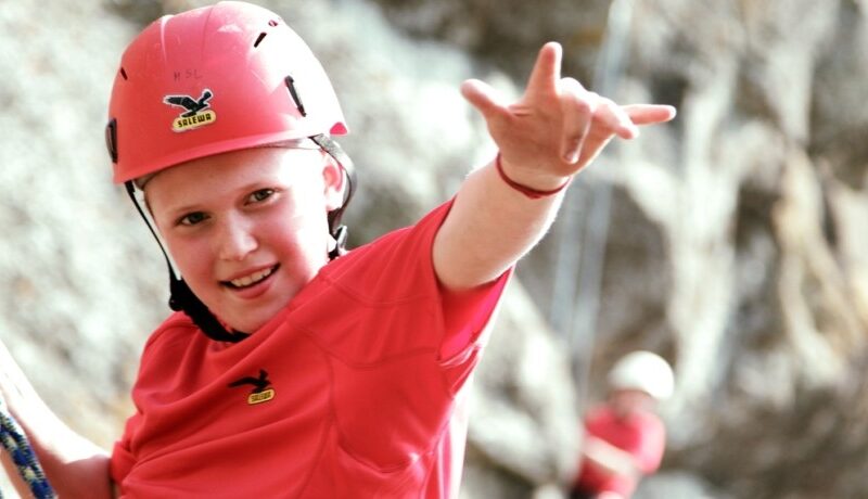 Ein Kind, das einen roten Helm und ein rotes Hemd trägt, lächelt und macht eine Geste mit einer Hand, während es im Freien klettert; eine andere Person ist unscharf im Hintergrund zu sehen.