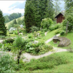 Ein Landschaftsgarten mit gewundenen Kieswegen, üppigem Grün, einer runden Holzkonstruktion und einem Holzhaus im Hintergrund inmitten von Bäumen und Hügeln.
