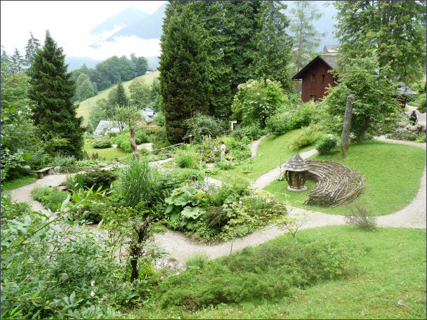 Ein Landschaftsgarten mit gewundenen Kieswegen, üppigem Grün, einer runden Holzkonstruktion und einem Holzhaus im Hintergrund inmitten von Bäumen und Hügeln.
