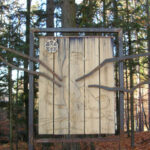 Ein Holzschild mit dem schwachen Umriss eines Drachens, umrahmt von Metallstäben, die die Flügel bilden, steht in einem Wald mit hohen Bäumen und herabgefallenen Blättern.