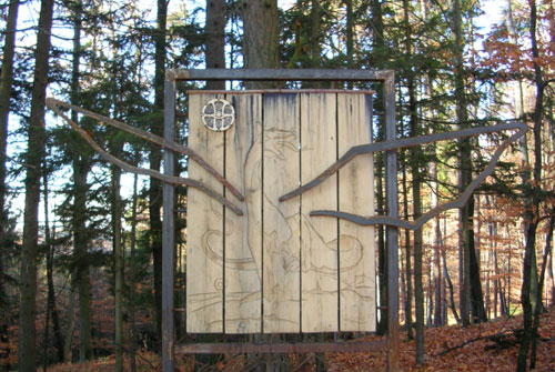 Ein Holzschild mit dem schwachen Umriss eines Drachens, umrahmt von Metallstäben, die die Flügel bilden, steht in einem Wald mit hohen Bäumen und herabgefallenen Blättern.