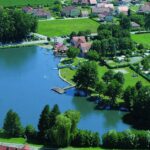 Luftaufnahme eines kleinen Sees mit einem Steg, einem grasbewachsenen Erholungsgebiet, geparkten Autos und Wohnhäusern im Hintergrund.