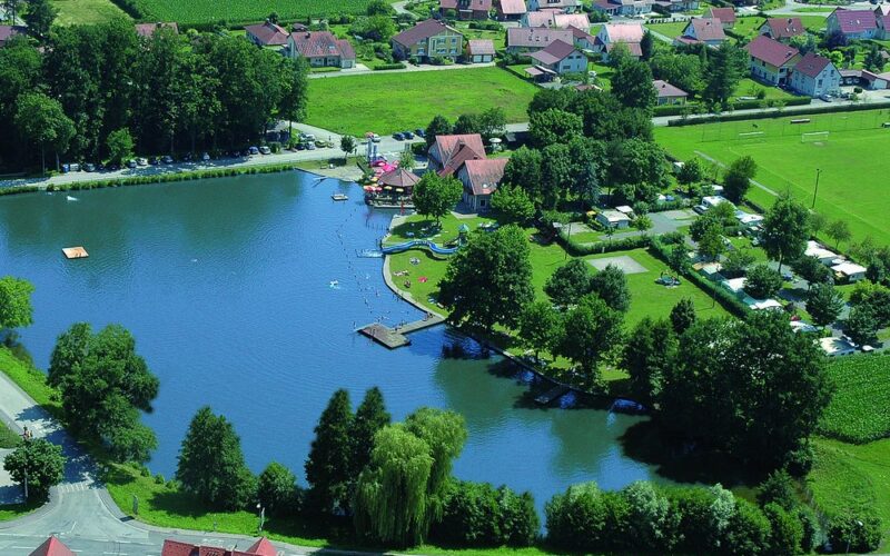 Luftaufnahme eines kleinen Sees mit einem Steg, einem grasbewachsenen Erholungsgebiet, geparkten Autos und Wohnhäusern im Hintergrund.