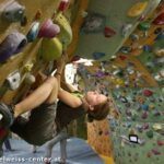 Eine Person klettert an einer überhängenden Boulderwand mit bunten Griffen in einer Kletterhalle.