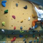 Ein Junge in einem weißen T-Shirt und einer orangefarbenen Hose klettert an einer Indoor-Kletterwand mit verschiedenfarbigen Griffen.