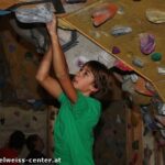 Eine Person in einem grünen Hemd klettert an einer Boulderwand in einer Halle und hält sich an einem Überhang fest. Im Hintergrund sind andere Kletterer zu sehen.