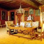 Ein historischer Speisesaal mit Holzfußboden, einem langen Tisch mit Porzellan, verzierten Stühlen, einem Kronleuchter und Porträts an den roten Wänden.