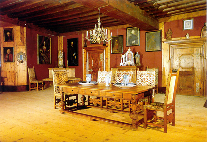 Ein historischer Speisesaal mit Holzfußboden, einem langen Tisch mit Porzellan, verzierten Stühlen, einem Kronleuchter und Porträts an den roten Wänden.