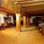 Ein historischer Raum mit Holzbalken und Säulen, in dem alte Möbel, dekorative Elemente und alte Geräte an den Wänden ausgestellt sind.