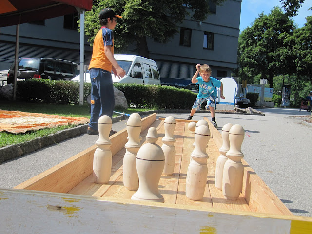 Ein Kind wirft eine Kugel, um große hölzerne Bowlingkegel auf einer hölzernen Außenbahn umzuwerfen, während ein Erwachsener in der Nähe steht.