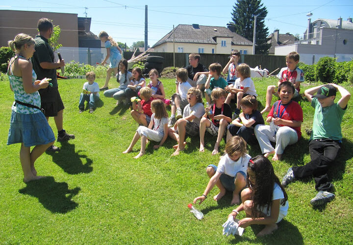 Eine Gruppe von Kindern sitzt und steht an einem sonnigen Tag auf einem Grashügel im Freien, im Hintergrund sind Häuser und Bäume zu sehen.