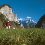 Ein kleines Kind in einer roten Jacke geht durch hohes Gras in einem Bergtal, mit felsigen Klippen und schneebedeckten Gipfeln im Hintergrund unter einem klaren blauen Himmel.