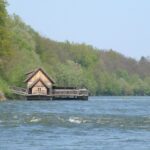 Ein hölzernes Bootshaus steht am Rande eines Flusses, umgeben von dichten grünen Bäumen unter einem klaren Himmel.