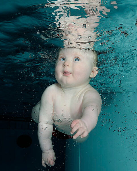 Ein Baby befindet sich in einem Schwimmbecken unter Wasser, schaut mit offenen Augen und ausgestreckten Armen nach oben und ist von Luftblasen umgeben.