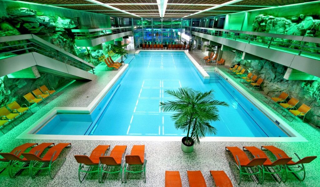 Hallenbad mit türkisfarbenem Wasser, umgeben von orangefarbenen Liegestühlen, Felswänden und grüner Beleuchtung, mit einer zentralen Palme und modernen architektonischen Elementen.