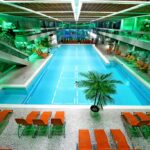 Hallenbad mit türkisfarbenem Wasser, umgeben von orangefarbenen Liegestühlen, Felswänden und grüner Beleuchtung, mit einer zentralen Palme und modernen architektonischen Elementen.