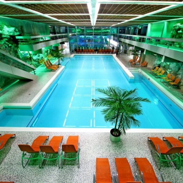 Hallenbad mit türkisfarbenem Wasser, umgeben von orangefarbenen Liegestühlen, Felswänden und grüner Beleuchtung, mit einer zentralen Palme und modernen architektonischen Elementen.