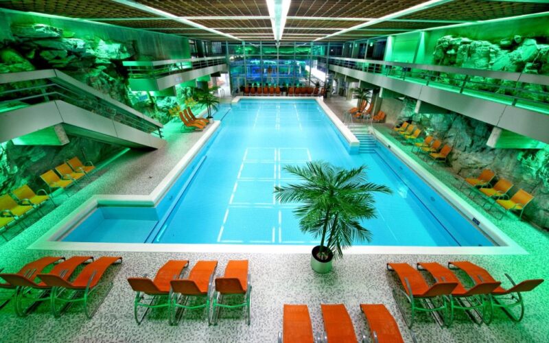 Hallenbad mit türkisfarbenem Wasser, umgeben von orangefarbenen Liegestühlen, Felswänden und grüner Beleuchtung, mit einer zentralen Palme und modernen architektonischen Elementen.