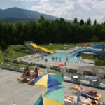 Die Menschen entspannen sich auf Liegestühlen in der Nähe eines Außenpools mit Wasserrutschen in einem grasbewachsenen Park, der von Bäumen und Bergen umgeben ist.