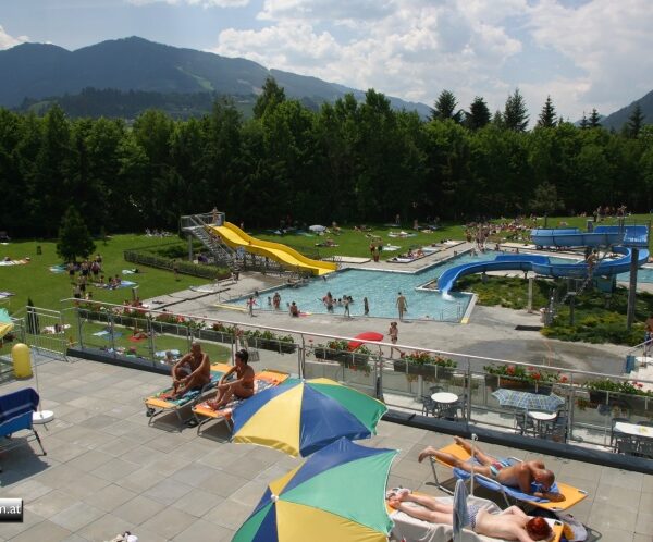 Die Menschen entspannen sich auf Liegestühlen in der Nähe eines Außenpools mit Wasserrutschen in einem grasbewachsenen Park, der von Bäumen und Bergen umgeben ist.