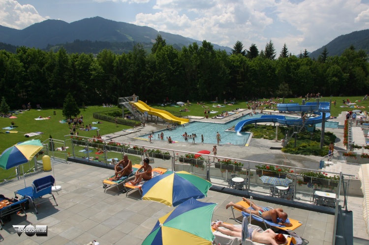 Die Menschen entspannen sich auf Liegestühlen in der Nähe eines Außenpools mit Wasserrutschen in einem grasbewachsenen Park, der von Bäumen und Bergen umgeben ist.