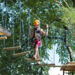Eine Person, die einen Helm und ein Gurtzeug trägt, überquert eine Seilbrücke in einem von grünen Bäumen umgebenen Outdoor-Abenteuerpark.