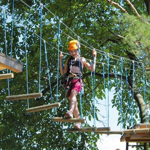 Eine Person, die einen Helm und ein Gurtzeug trägt, überquert eine Seilbrücke in einem von grünen Bäumen umgebenen Outdoor-Abenteuerpark.